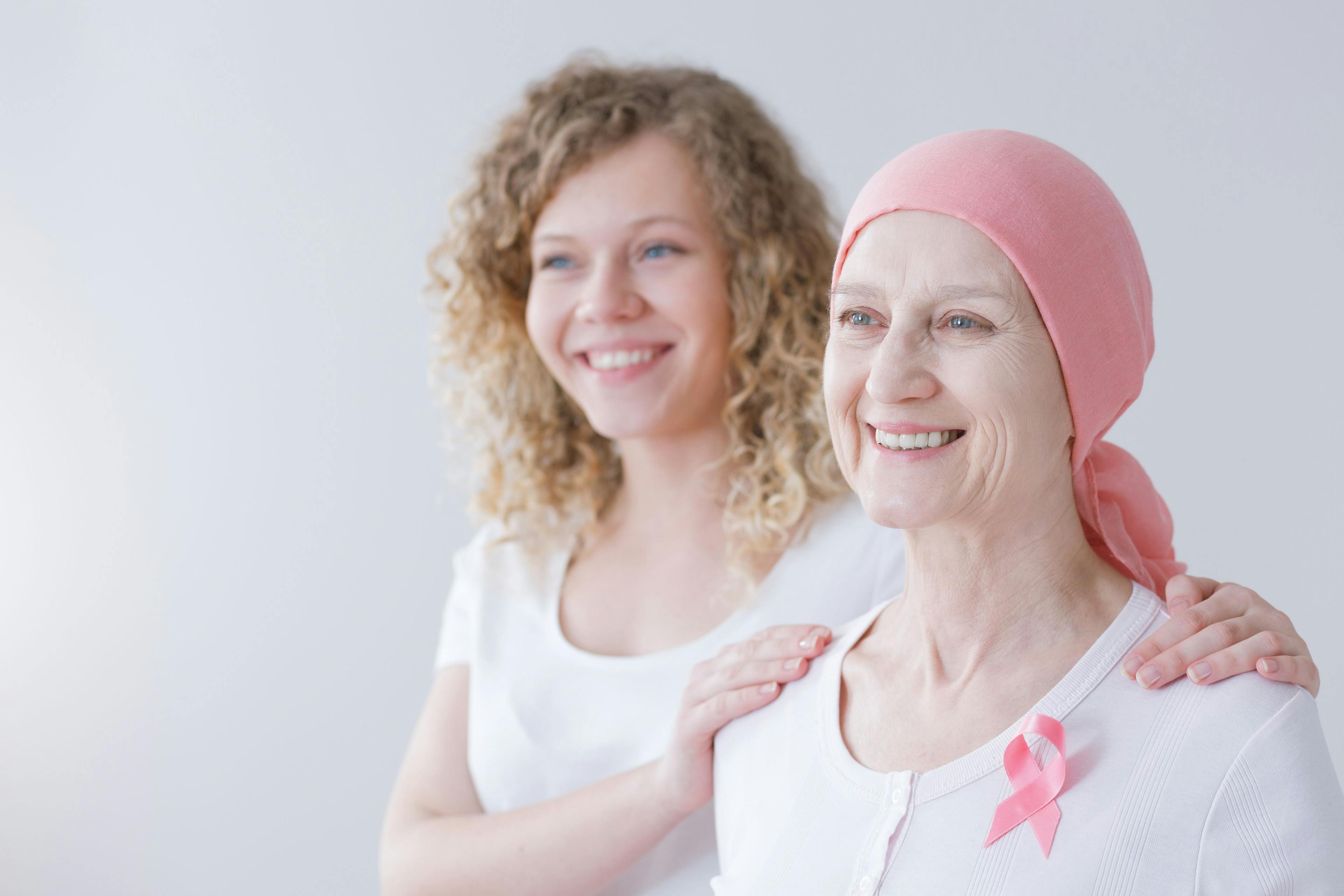 a young woman putting her hands on another woman's shoulders who has breast cancer and is wearing a pink ribbon and head scarf